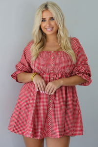 A woman wearing a red and white gingham mini dress with puff, elbow length sleeves and button detailing along the front, standing indoors against a white wall.