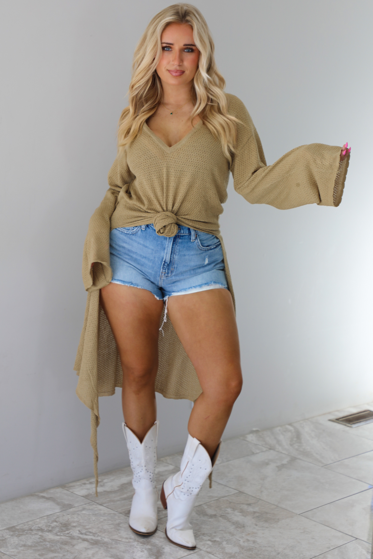 Woman wearing a camel long-sleeve top that's tied in the front with a knot and has a train in the back. The top is paired with denim shorts and white cowboy boots. The photo is taken indoors against a white wall with white marble flooring. 