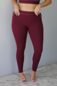 A woman wearing a maroon matching two piece set featuring a sports bra and high waisted leggings with white stitching details, standing indoors against a white wall with white marble flooring. The photo shows a closeup of the pants only. 