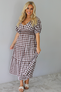 A woman modeling a brown and white gingham tiered midi dress with puff sleeves and a smocked waist, paired with beige slide sandals, standing indoors against a white wall with white marble flooring. 