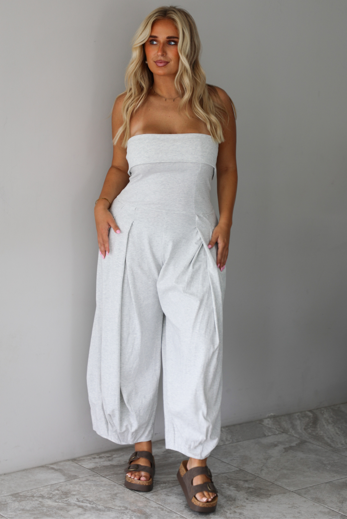 A woman modeling a light gray strapless jumpsuit with wide, balloon-style legs and gathered detailing, standing indoors against a white wall with white marble flooring. 