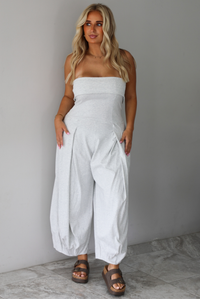 A woman modeling a light gray strapless jumpsuit with wide, balloon-style legs and gathered detailing, standing indoors against a white wall with white marble flooring. 