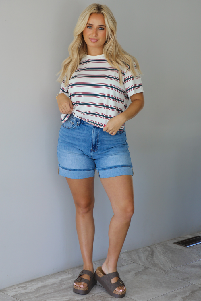 Woman wearing a striped, slightly cropped tee with light-wash denim shorts indoors against a white wall with white marble flooring. 