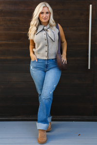 Woman model wearing a taupe sweater vest with black stitched detailing, has a tie detail at the neckline, is sleeveless, has functional button closures in the front, and has a ribbed texture. The sweater vest is paired with a medium wash denim, a dark brown leather purse, and ugg boots. The photo is taken outdoors against a dark brown wooden door. 