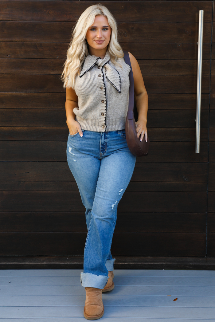 Woman model wearing a taupe sweater vest with black stitched detailing, has a tie detail at the neckline, is sleeveless, has functional button closures in the front, and has a ribbed texture. The sweater vest is paired with a medium wash denim, a dark brown leather purse, and ugg boots. The photo is taken outdoors against a dark brown wooden door. 