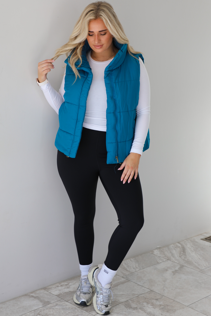 Woman wearing a teal puffer vest, white long-sleeve shirt, black leggings, and gray sneakers with white ankle socks indoors against a white wall with white marble flooring.
