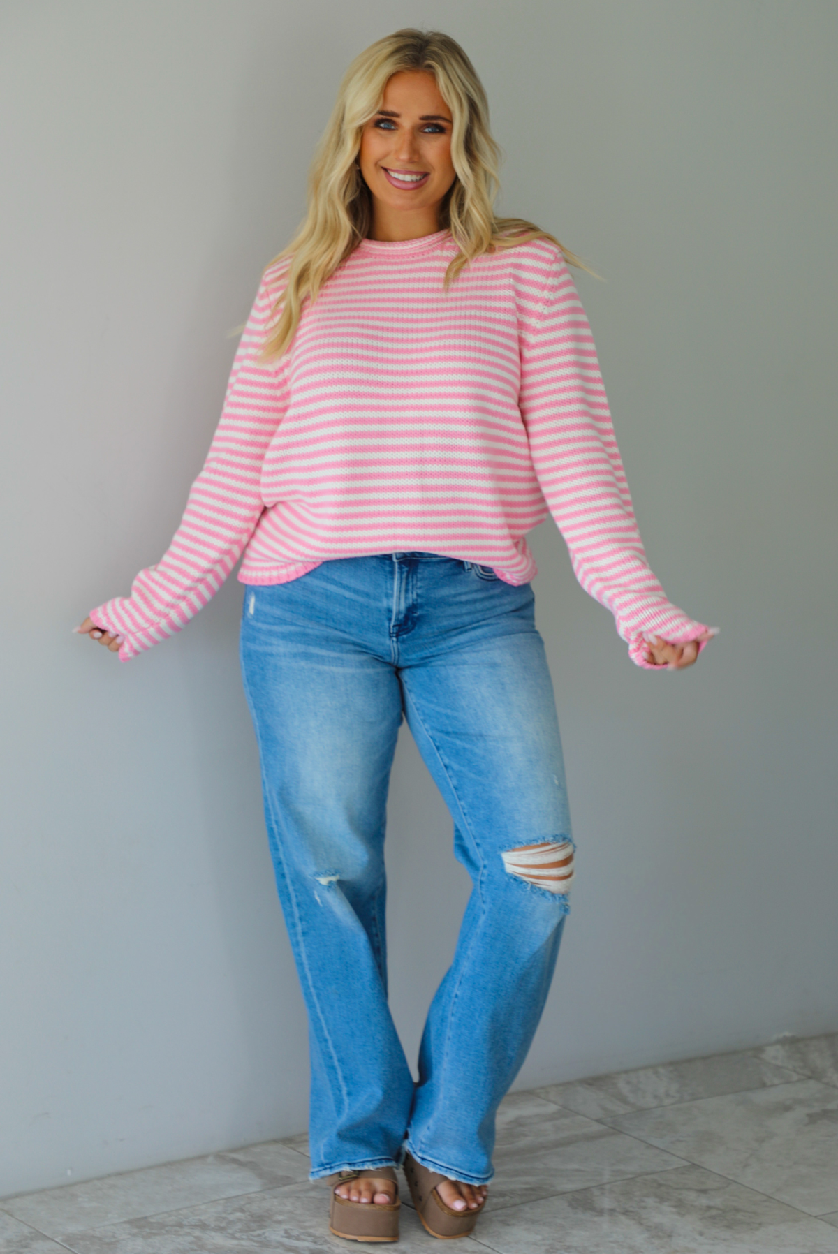 A woman wearing a pink and white striped sweater paired with light wash denim jeans, standing indoors against a white wall with white marble flooring. 