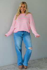 A woman wearing a pink and white striped sweater paired with light wash denim jeans, standing indoors against a white wall with white marble flooring. 