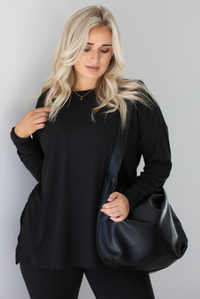 Woman wearing a black two piece set that features an oversized long sleeved top and fitted leggings. The set is paired with a black vegan leather shoulder bag. The photo shows a closeup of the top and bag. The photo is taken indoors against a white wall with white marble flooring.