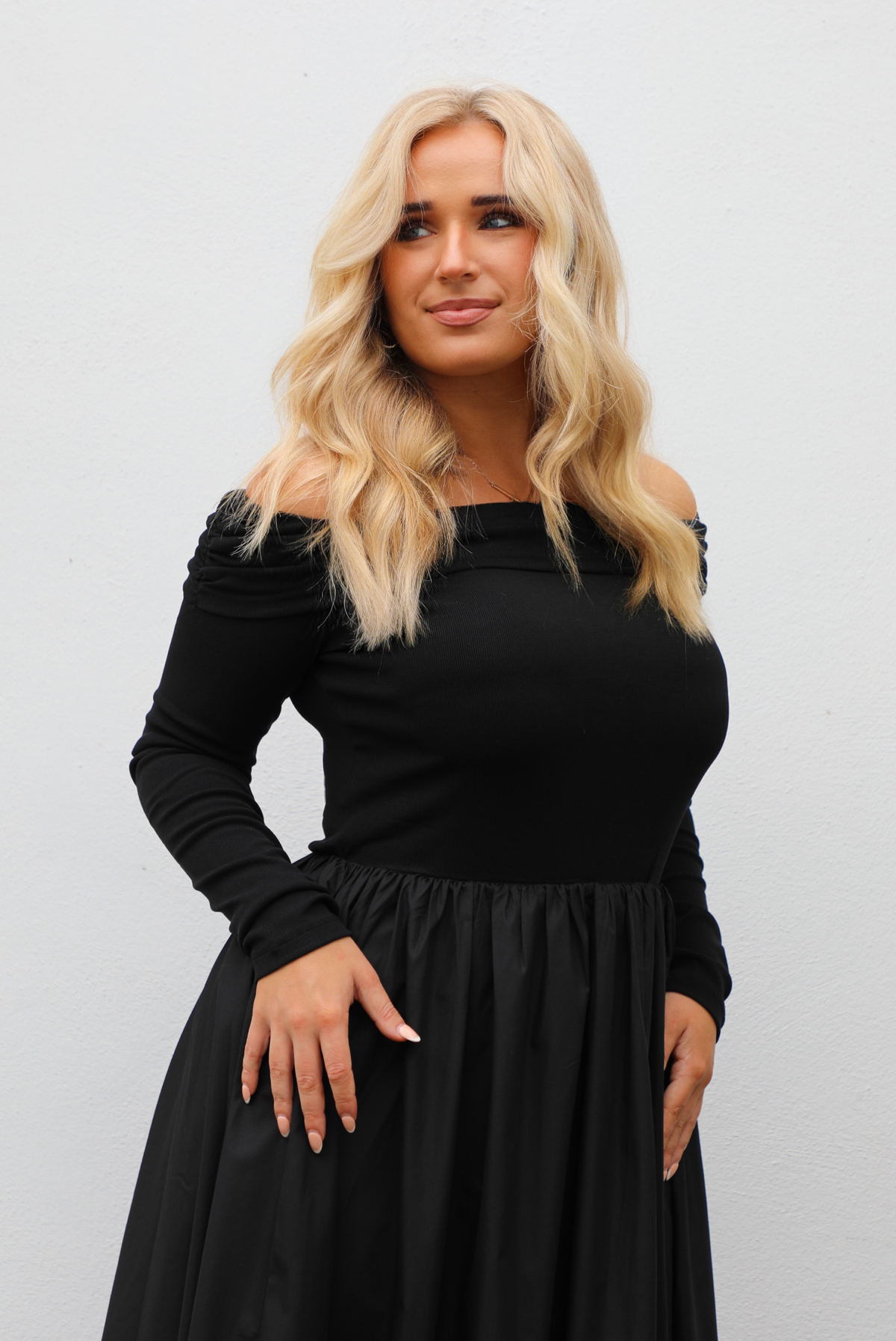Woman wearing a black off the shoulder, long sleeved midi length dress with a pair of clear strapped nude heels. The photo shows a close up of the bodice and upper skirt of the dress and the difference in textures. The bodice is fitted with a folded over detail at the upper arm, and the skirt is flowy. The photo is taken outdoors against a white wall.