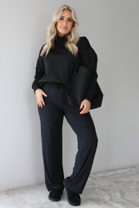 Woman wearing a black high-neck, oversized sweater, high waisted black pants, with a black shoulder bag, and black ugg boot shoes. The photo is taken indoors against a white wall with white marbled flooring. 