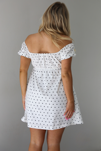 Woman wearing a white dress with a black polka dot print throughout. The photo is taken indoors against a white wall. The photo shows the back of the dress. 