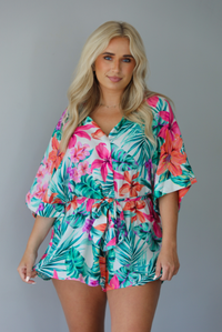 Woman wearing a multi-colored tropical romper with a collared neckline, flowy short sleeves, a cinched with a tie waistline, and flowy shorts indoors against a white wall.