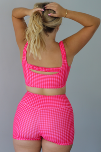 A woman wearing a red and hot pink gingham two-piece activewear set consisting of a crop top and matching shorts, standing indoors against a white wall. The photo shows the back of the set. 