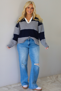 A woman wearing a navy and charcoal boyfriend style pullover top with a white collared detail, with distressed denim, standing indoors against a white wall with white marble flooring. 