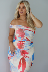A woman modeling a strapless off-the-shoulder maxi dress with a vibrant red, blue, and cream floral print against a white wall. The photo shows a closeup of the off the shoulder detail. 