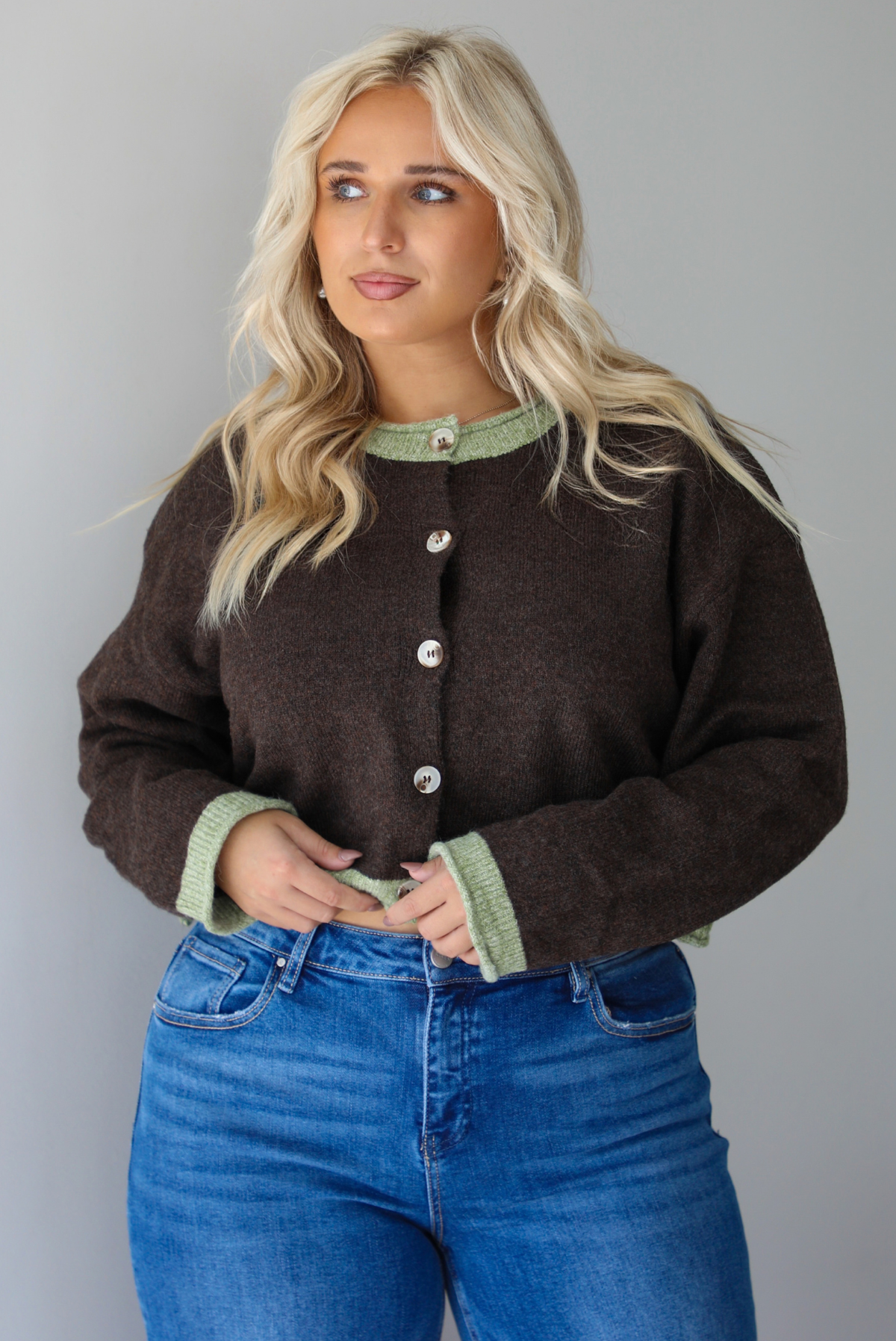 A woman wearing a dark brown with green trim cardigan fully buttoned up paired with dark wash denim jeans, standing indoors against a white wall.