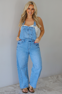 Woman wearing a pair of denim overalls with a white floral strapless top underneath indoors against a white wall with white marble flooring. 