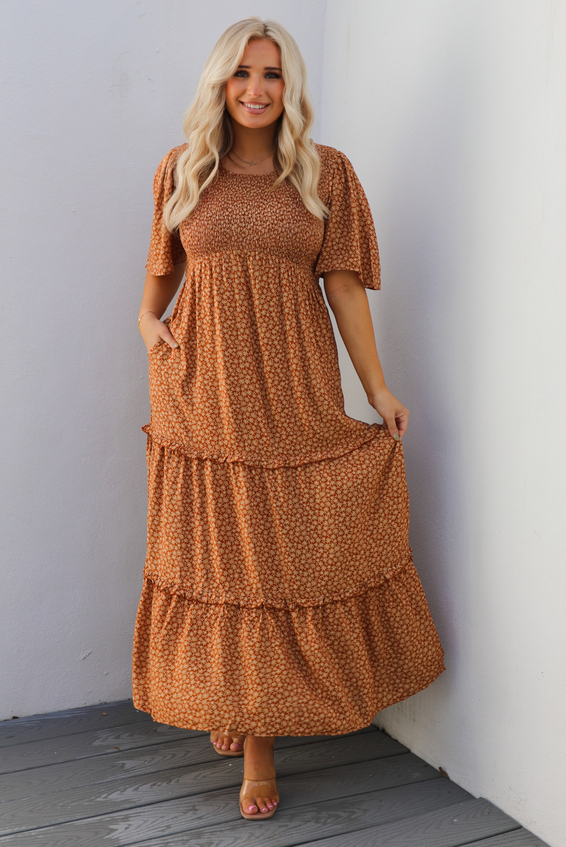 A woman model outdoors wearing a rust dress with a cream floral print pattern throughout. It has flowy short sleeves, a smocked bodice, and a flowy maxi length skirt with functional pockets on either side. 