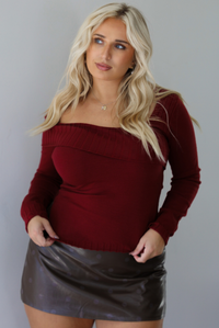Woman wearing a maroon knit, long sleeved, off the shoulder knit top paired with a dark brown vegan leather mini skirt with a monochromatic polka dot print pattern, standing indoors against a white wall.