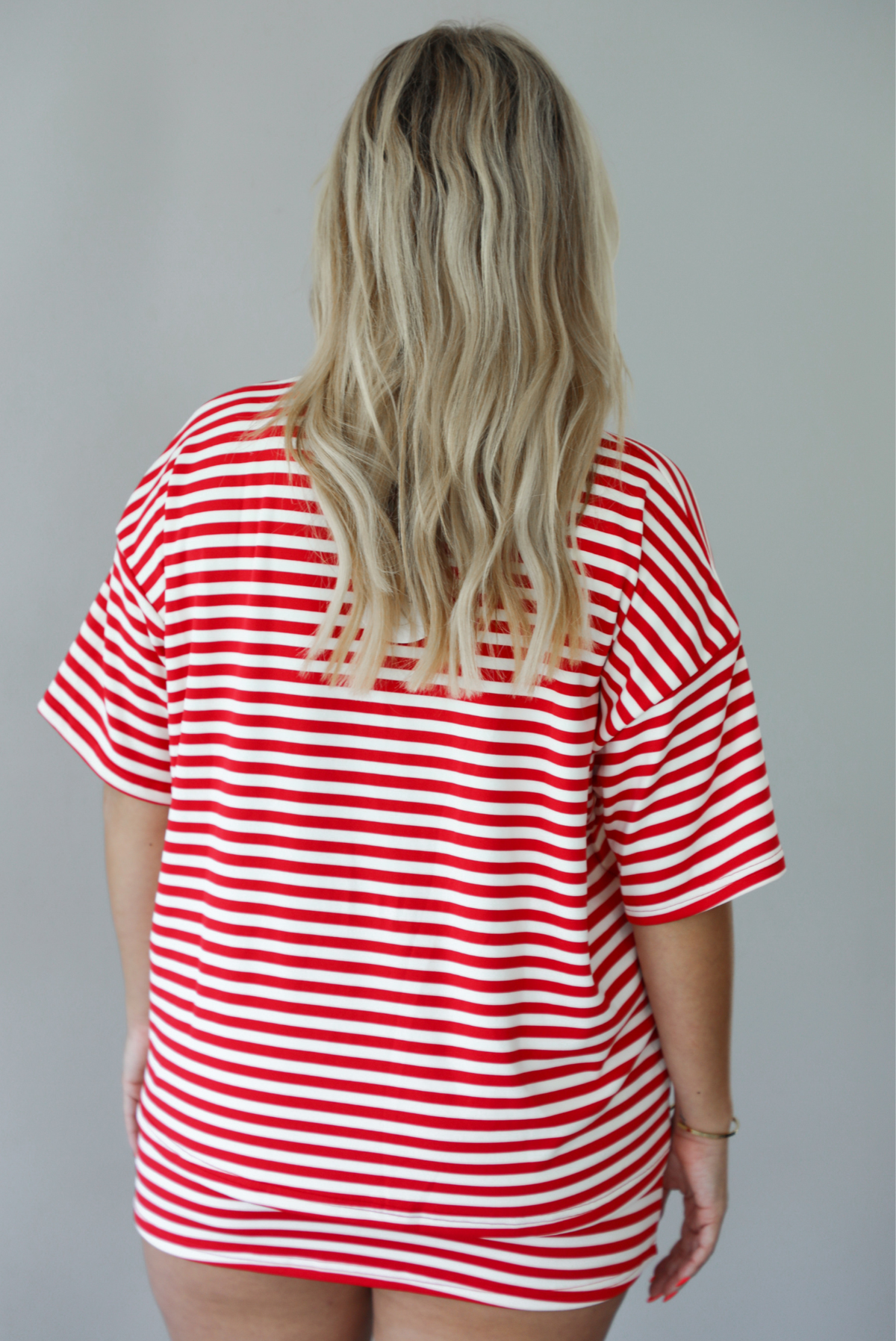 Woman wearing a red and white striped set featuring an oversized top and mini skirt indoors against a white wall. The photo shows the back of the set. 