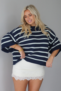 A woman wearing a navy and multi-colored striped design knit top paired with a fitted cream, lace trim mini skirt, standing indoors against a white wall.