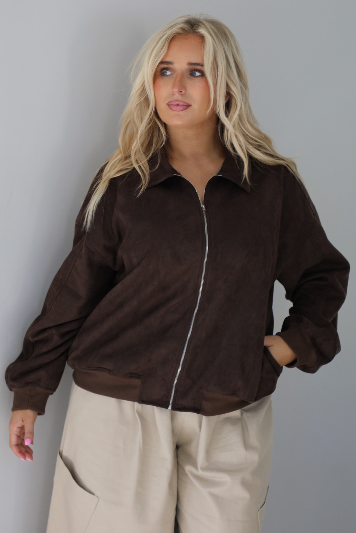 A woman wearing a brown suede jacket with a white tank and sand colored baggy pants, standing indoors against a white wall. The photo shows a closeup of the jacket, which is zipped closured, and the models hand is in one of the side pockets. 