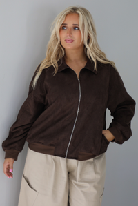 A woman wearing a brown suede jacket with a white tank and sand colored baggy pants, standing indoors against a white wall. The photo shows a closeup of the jacket, which is zipped closured, and the models hand is in one of the side pockets. 