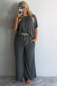 A woman wearing a charcoal and white striped two piece matching set with green trim detailing at the neckline and waistline, featuring an oversized short sleeved top and high waisted pants, standing indoors taking a mirror selfie against a white wall with white marble flooring. 