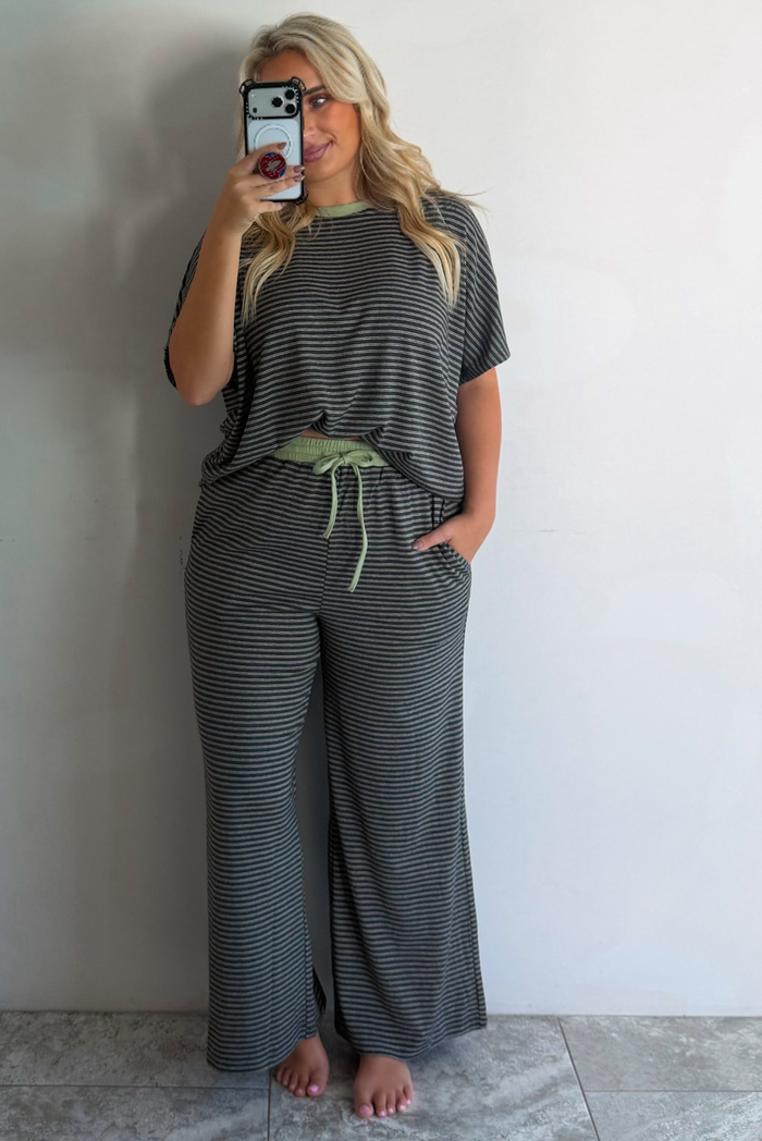 A woman wearing a charcoal and white striped two piece matching set with green trim detailing at the neckline and waistline, featuring an oversized short sleeved top and high waisted pants, standing indoors taking a mirror selfie against a white wall with white marble flooring. 