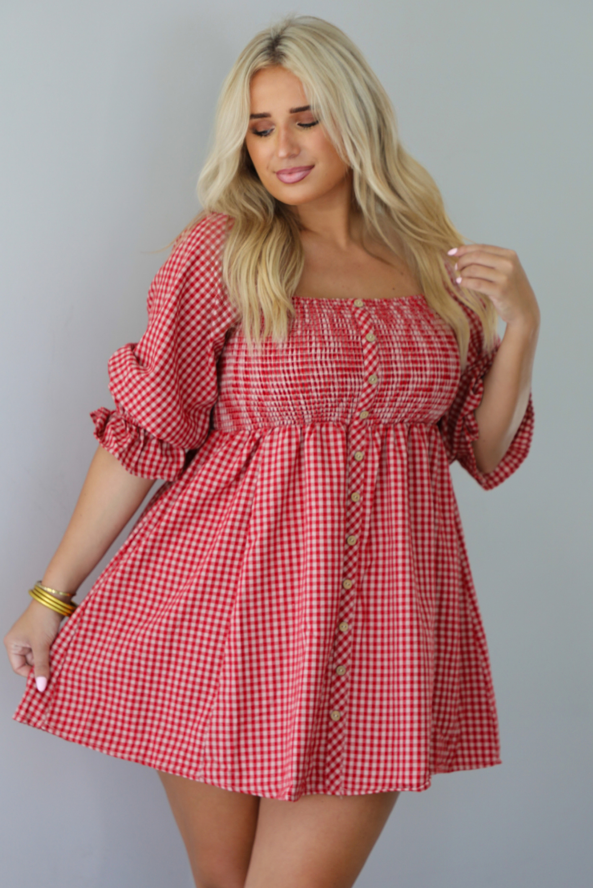 A woman wearing a red and white gingham mini dress with puff, elbow length sleeves and button detailing along the front, standing indoors against a white wall.