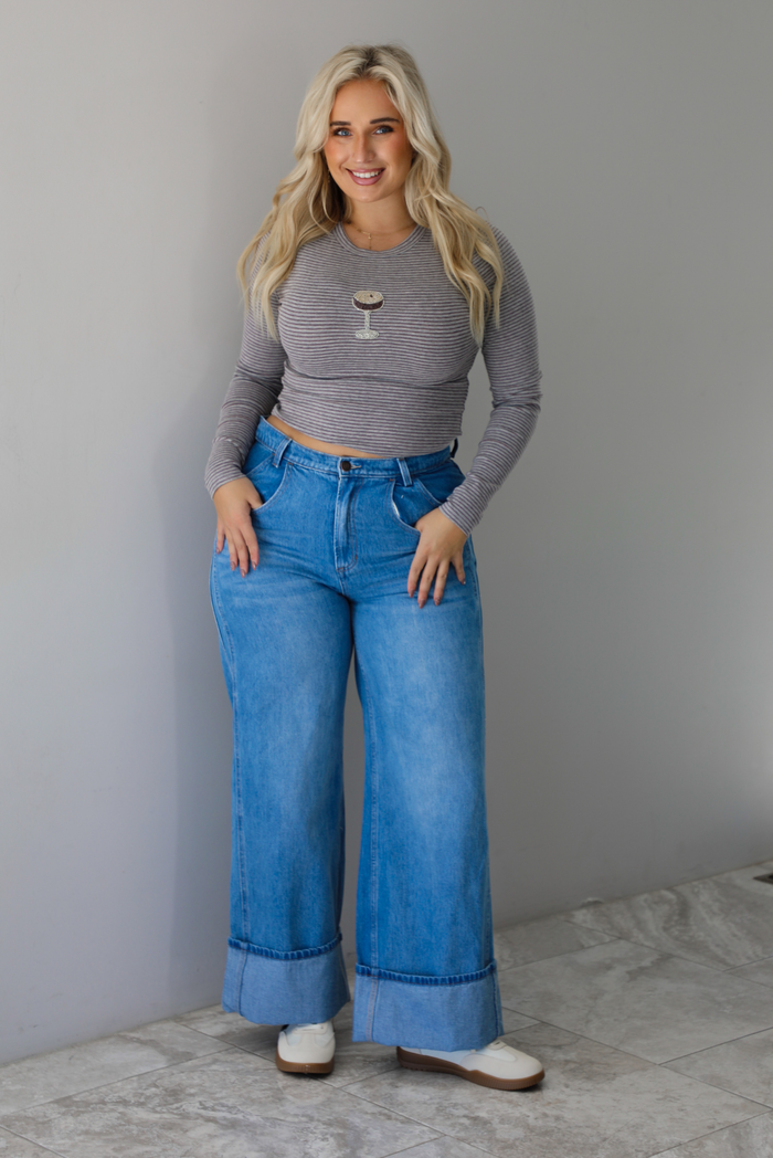 A woman wearing a striped, cropped long sleeved top with a beaded espresso martini on the front with medium wash denim jeans, standing indoors against a white wall with white marble flooring 