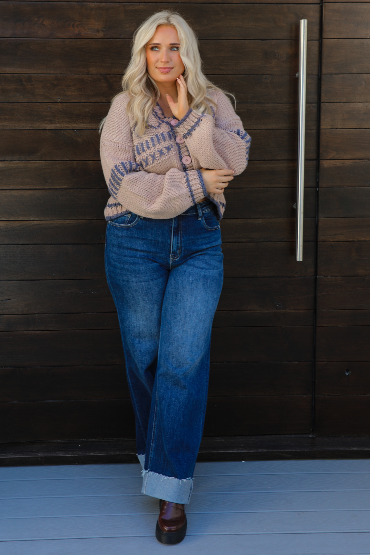 Woman model wearing a mocha brown cardigan that has grey stitching details throughout. The cardigan has a cropped cut, is oversized, and has functional button closures in the front. She's wearing the cardigan with dark denim jeans and brown shoes. The photo is taken against a dark brown wooden door outdoors. 