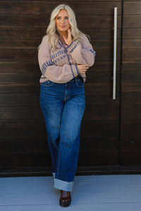 Woman model wearing a mocha brown cardigan that has grey stitching details throughout. The cardigan has a cropped cut, is oversized, and has functional button closures in the front. She's wearing the cardigan with dark denim jeans and brown shoes. The photo is taken against a dark brown wooden door outdoors. 