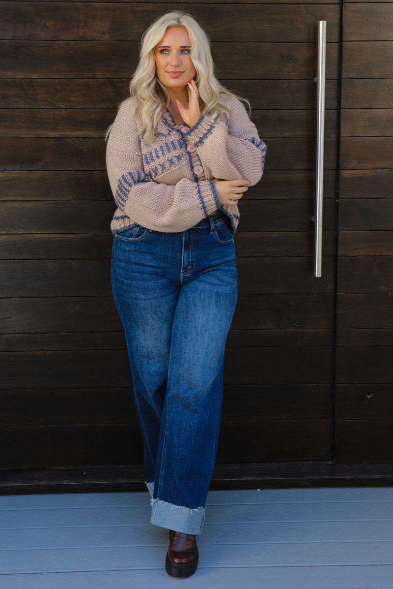 Woman model wearing a mocha brown cardigan that has grey stitching details throughout. The cardigan has a cropped cut, is oversized, and has functional button closures in the front. She's wearing the cardigan with dark denim jeans and brown shoes. The photo is taken against a dark brown wooden door outdoors. 