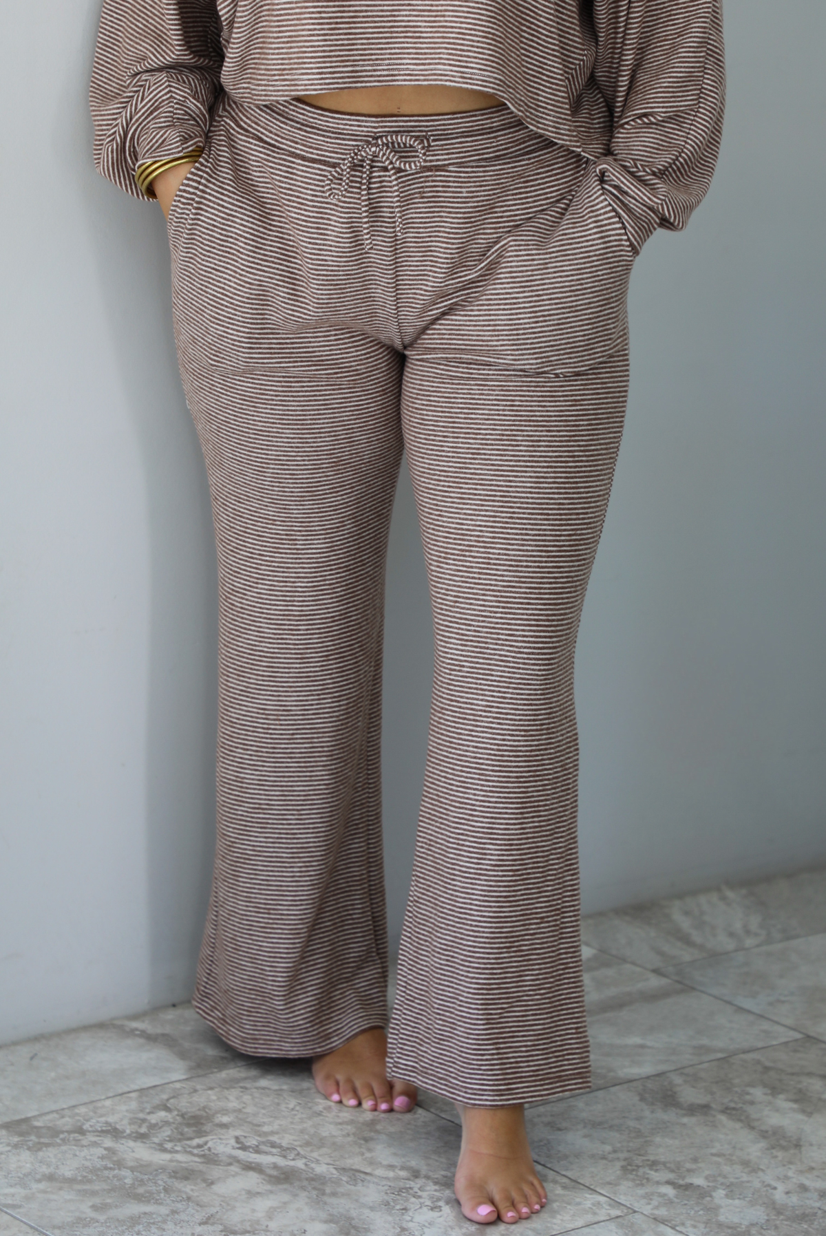 A woman wearing a mocha and white striped matching two piece set featuring a cropped, flowy. long sleeved top paired with high waisted wide leg pants, standing indoors against a white wall with white marble flooring. The photo shows a closeup of the pants, with the model having their hands in the pockets on either side, with an elastic and drawstring waistline shown. 