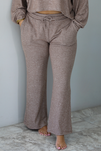 A woman wearing a mocha and white striped matching two piece set featuring a cropped, flowy. long sleeved top paired with high waisted wide leg pants, standing indoors against a white wall with white marble flooring. The photo shows a closeup of the pants, with the model having their hands in the pockets on either side, with an elastic and drawstring waistline shown. 