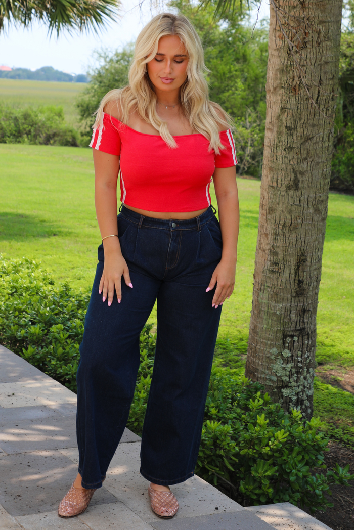 A woman standing outdoors on a stone walkway wearing a bright red off-the-shoulder crop top with white sleeve stripes, dark wide-leg denim jeans, and clear woven sandals, with a grassy yard and trees in the background.