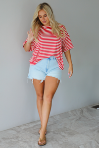 A woman modeling a red and white horizontally striped short-sleeve t-shirt with light blue denim shorts and brown sandals, standing indoors against a white wall with white marble flooring. 