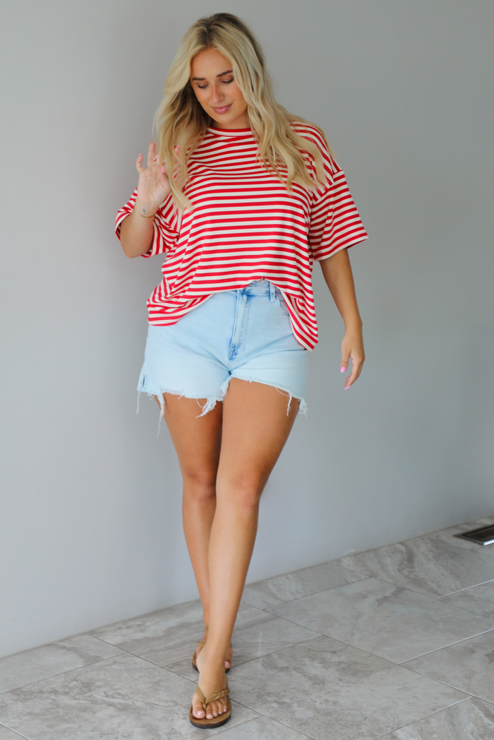 A woman modeling a red and white horizontally striped short-sleeve t-shirt with light blue denim shorts and brown sandals, standing indoors against a white wall with white marble flooring. 