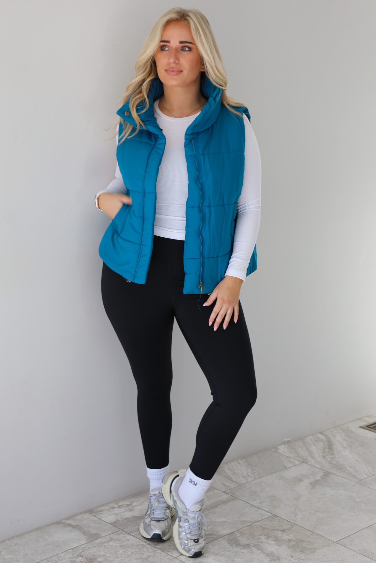 Woman wearing a teal puffer vest, white long-sleeve shirt, black leggings, and gray sneakers with white ankle socks indoors against a white wall with white marble flooring.