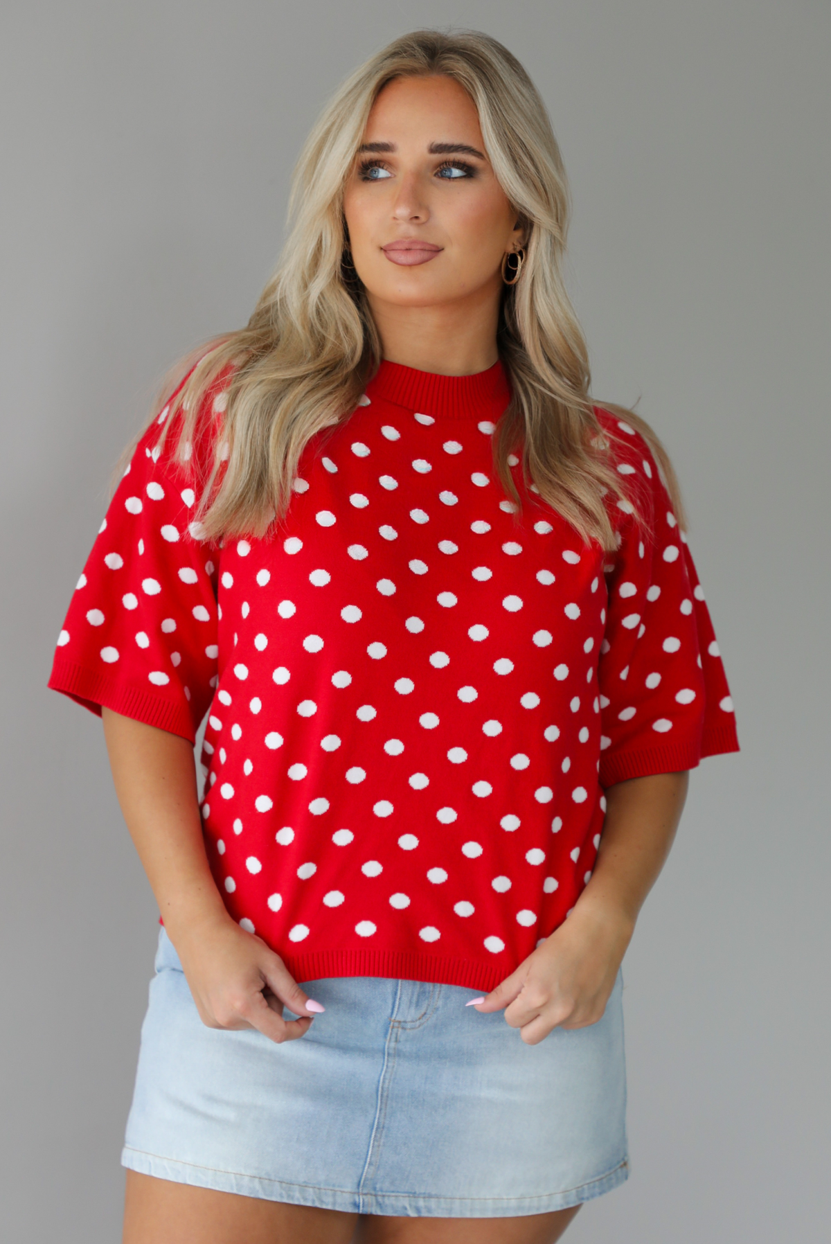 Woman wearing a red knit top with a white polka dot print pattern throughout with a high waisted, light-wash denim skort. The photo is taken indoors against a white wall.