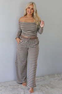 A woman wearing a brown with cream striped matching two piece set consisting of a long sleeved, off the shoulder top and high waisted pants, standing indoors against a white wall with white marble flooring. 