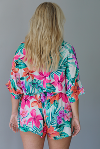 Woman wearing a multi-colored tropical romper with a collared neckline, flowy short sleeves, a cinched with a tie waistline, and flowy shorts indoors against a white wall. The photo shows the back of the romper. 