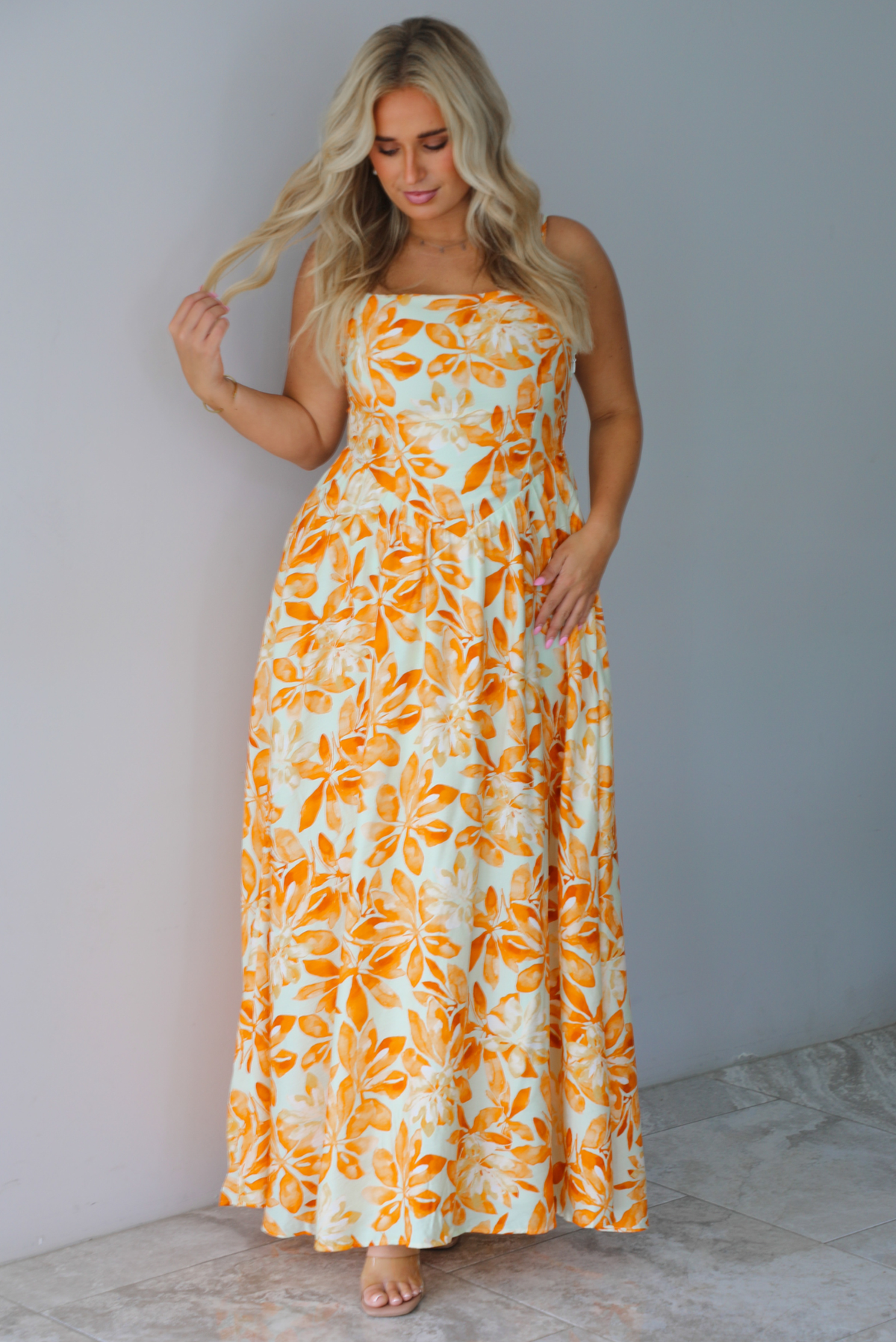 A woman modeling a spaghetti strapped, floor-length maxi dress with a white base and a vibrant marigold orange floral pattern, standing indoors against a white wall with white marble flooring. 
