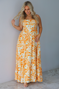 A woman modeling a spaghetti strapped, floor-length maxi dress with a white base and a vibrant marigold orange floral pattern, standing indoors against a white wall with white marble flooring. 