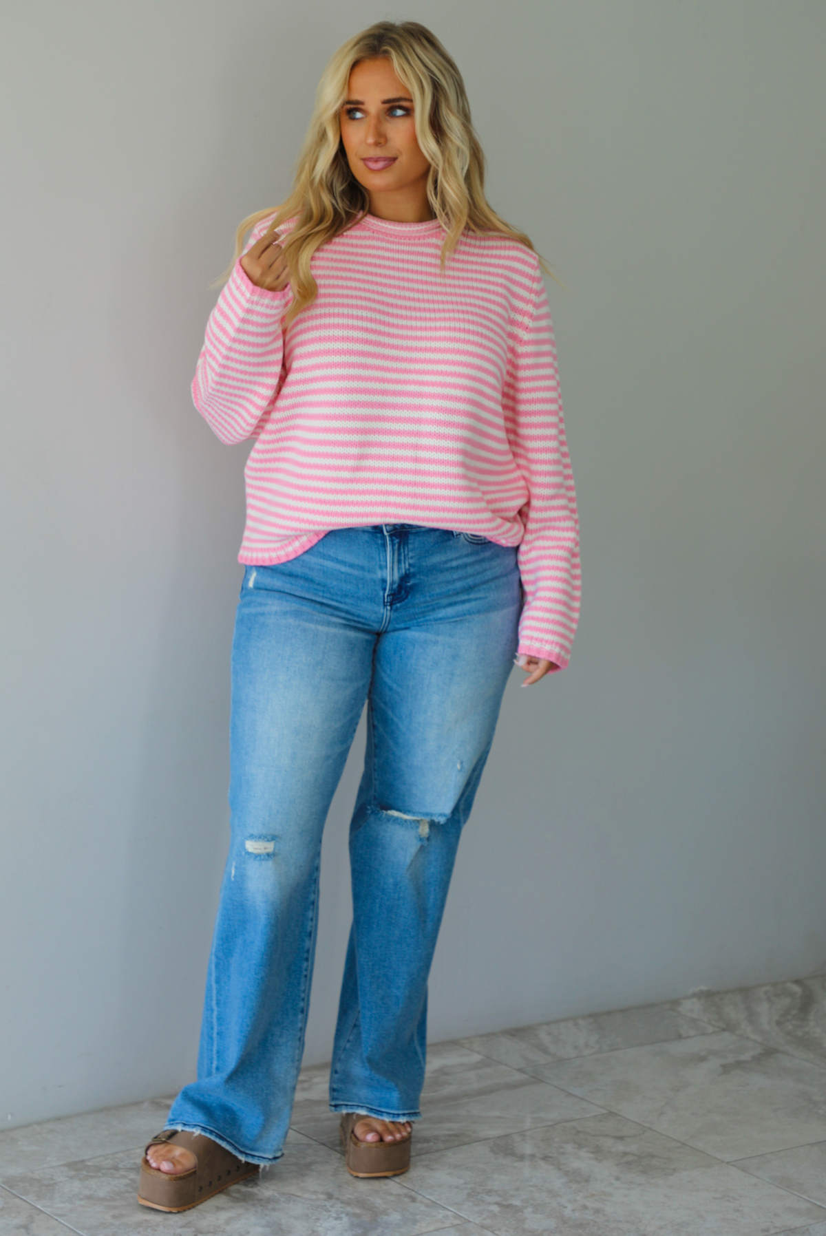 A woman wearing a pink and white striped sweater paired with light wash denim jeans, standing indoors against a white wall with white marble flooring. 