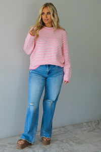 A woman wearing a pink and white striped sweater paired with light wash denim jeans, standing indoors against a white wall with white marble flooring. 