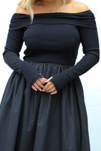 Woman wearing a black off the shoulder, long sleeved midi length dress with a pair of clear strapped nude heels. The photo shows a close up of the bodice and upper skirt of the dress and the difference in textures. The bodice is fitted with a folded over detail at the upper arm, the skirt is flowy, and the sleeves have thumbhole cut outs. The photo is taken outdoors against a white wall.