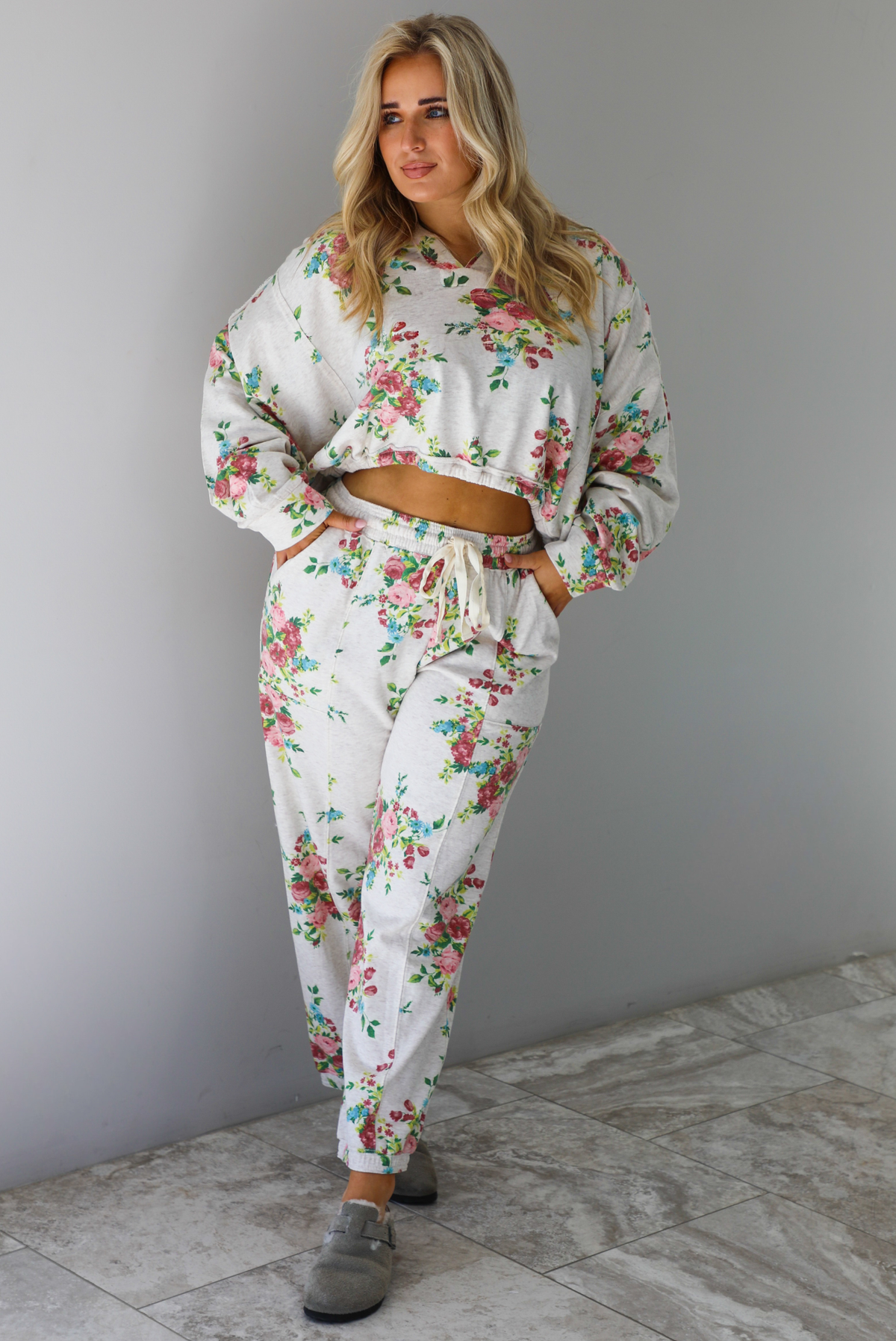 Woman wearing an oatmeal colored and multi-colored floral print two piece set featuring a collared long sleeved crop top and high waisted pants. The photo is taken indoors against a white wall with white marble flooring. 